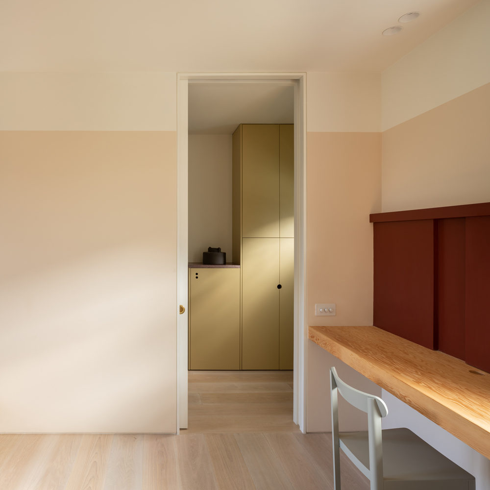 Light-filled study with a long timber desk, warm peach-toned walls and a view through to mustard-coloured storage cabinets.