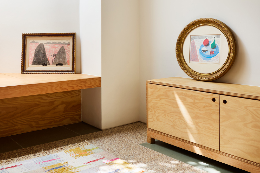 Framed works displayed on plywood furniture in a sunlit corner.
