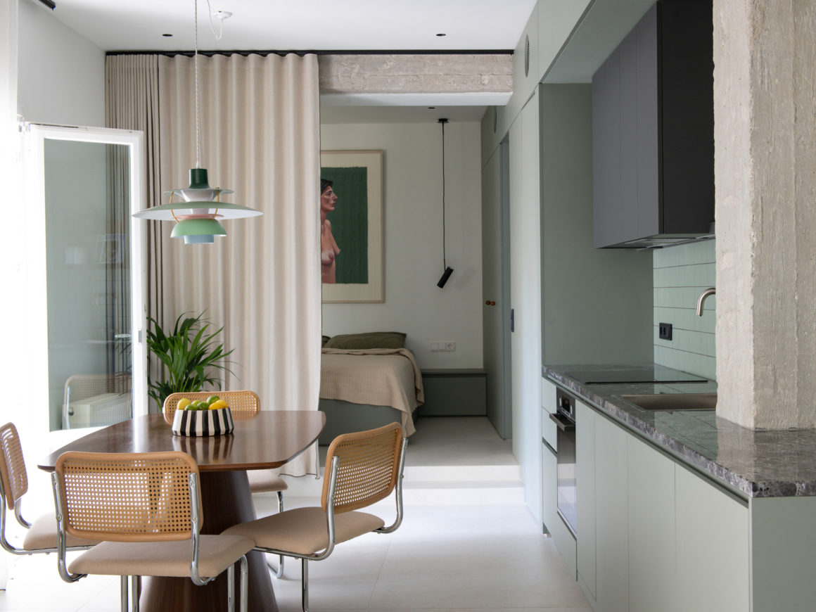 Open-plan living room with green cabinetry, timber flooring and natural light filtering through large windows.
